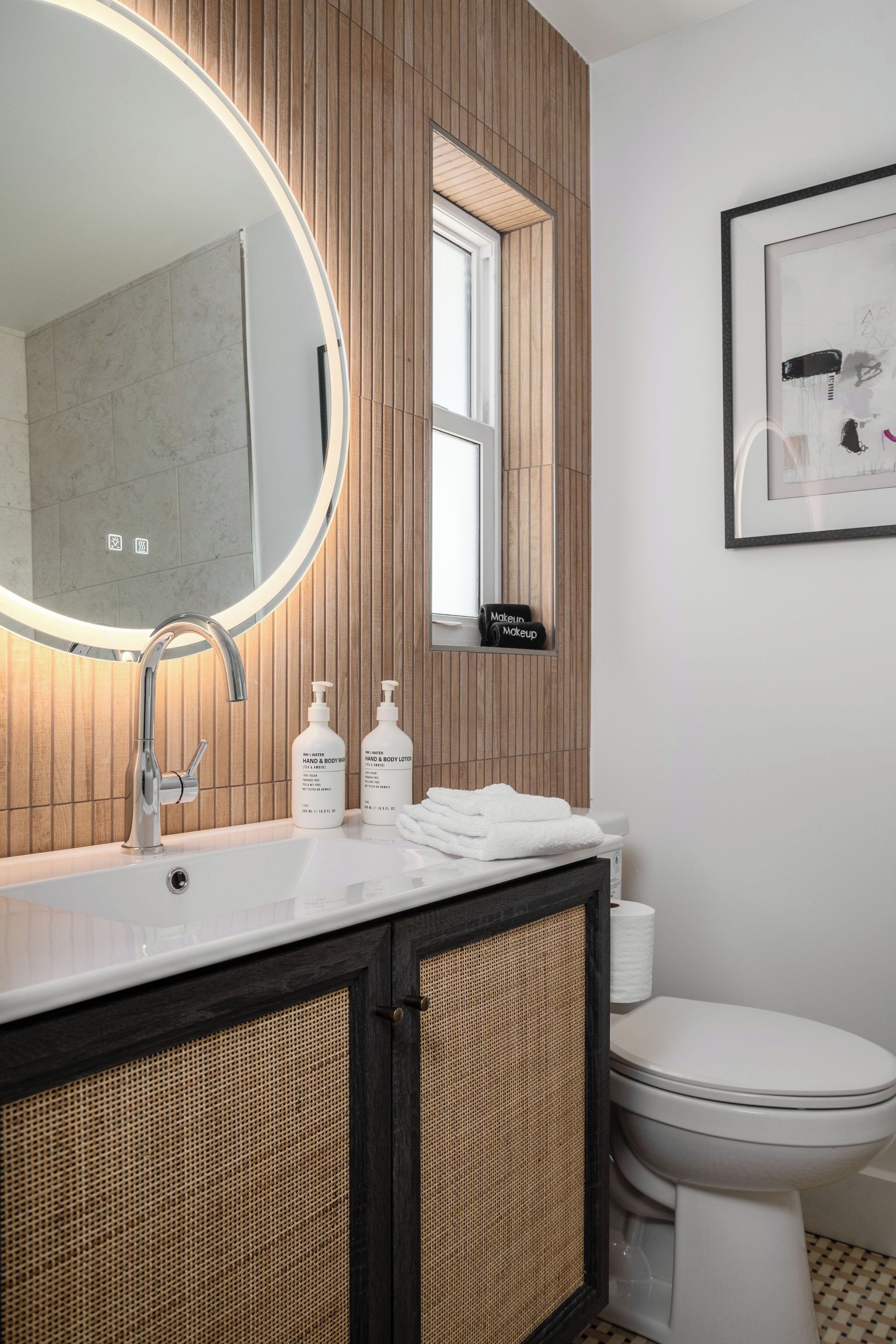 A bathroom with a round mirror, a wooden vanity, a toilet, and a framed artwork on the wall.