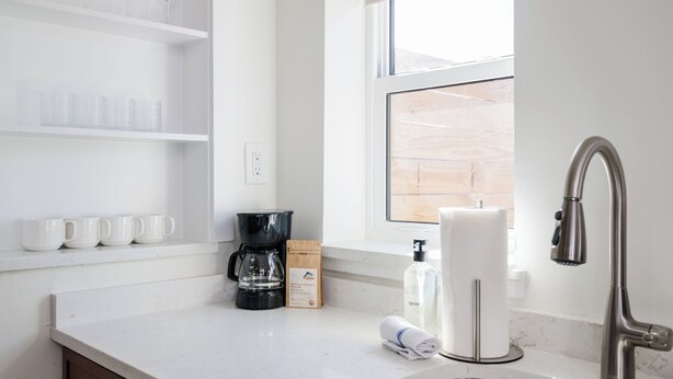 Una cocina moderna con encimeras blancas, un lavamanos de acero inoxidable, una cafetera y una ventana con vistas al exterior.