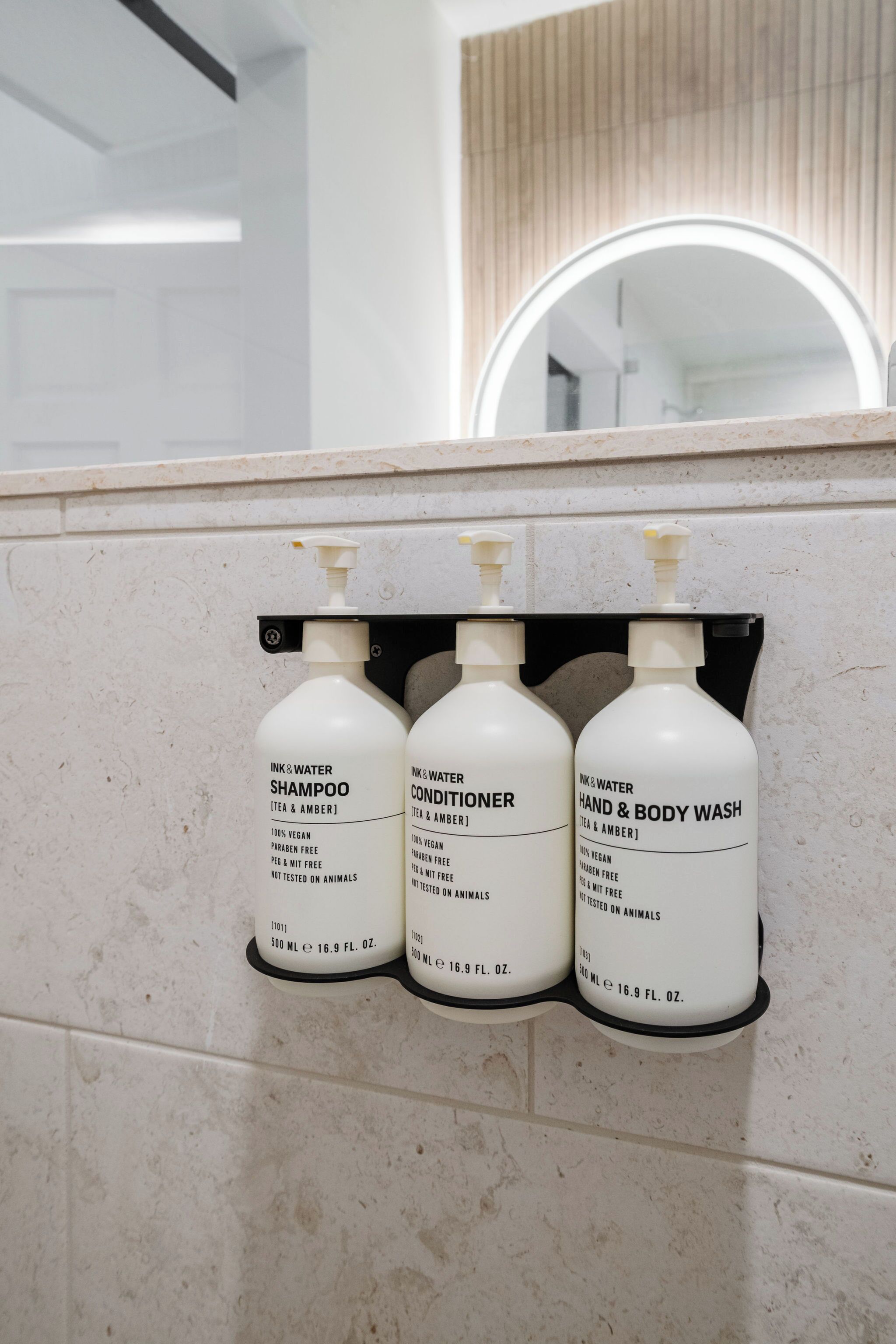 A bathroom with three bottles of shampoo, conditioner, and hand & body wash on a wall-mounted dispenser.
