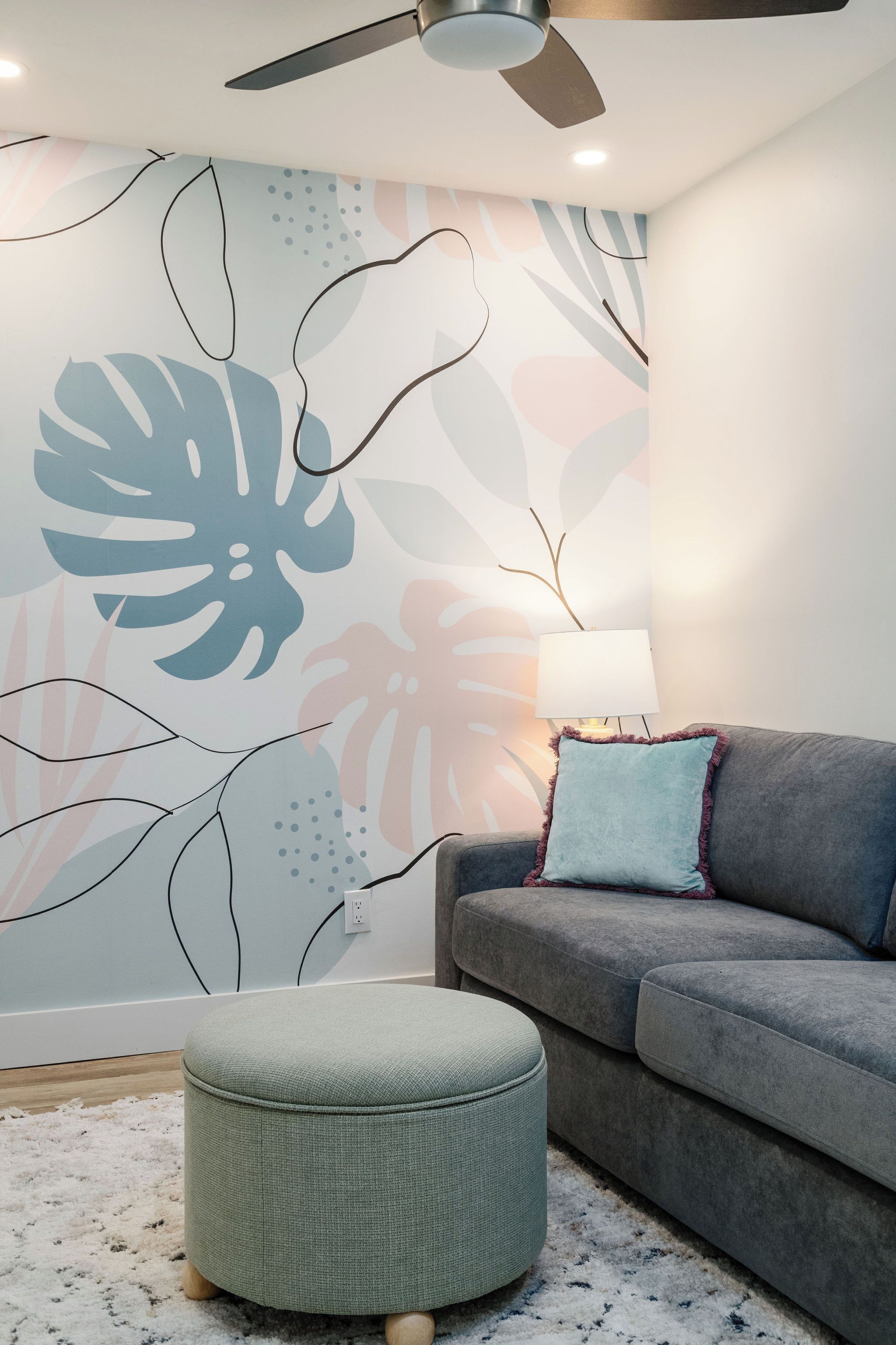 A living room with a grey sofa, a round ottoman, a ceiling fan, and a wall with abstract leaf patterns.