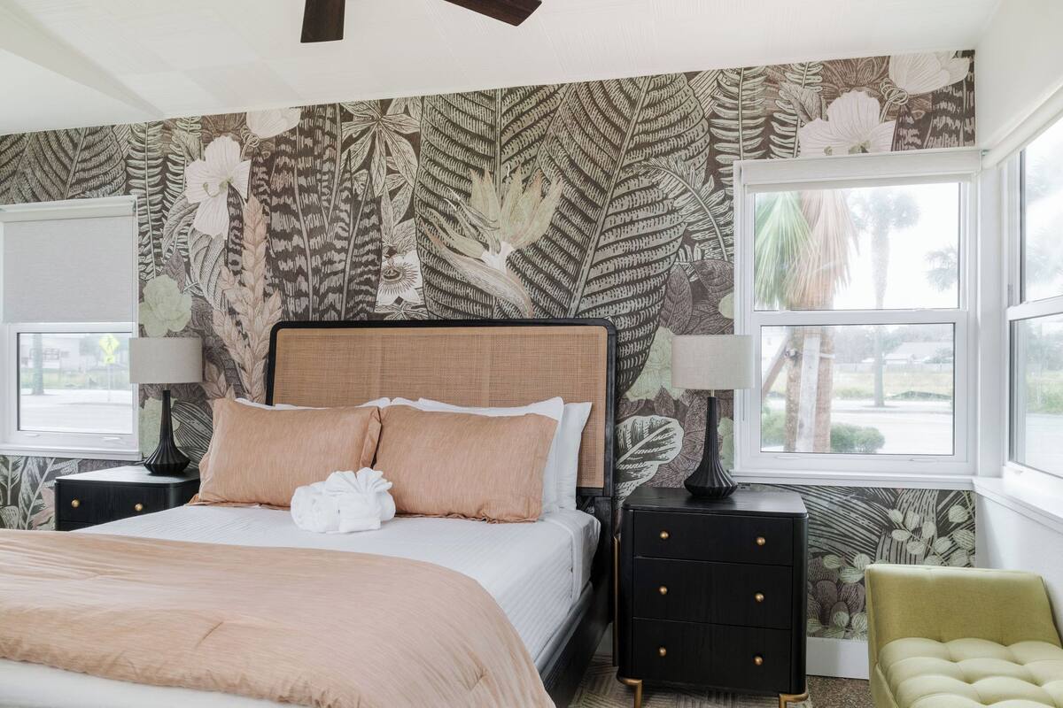 Un dormitorio con una cama grande, dos mesitas de noche, un sillón verde y una ventana con vista a palmeras.