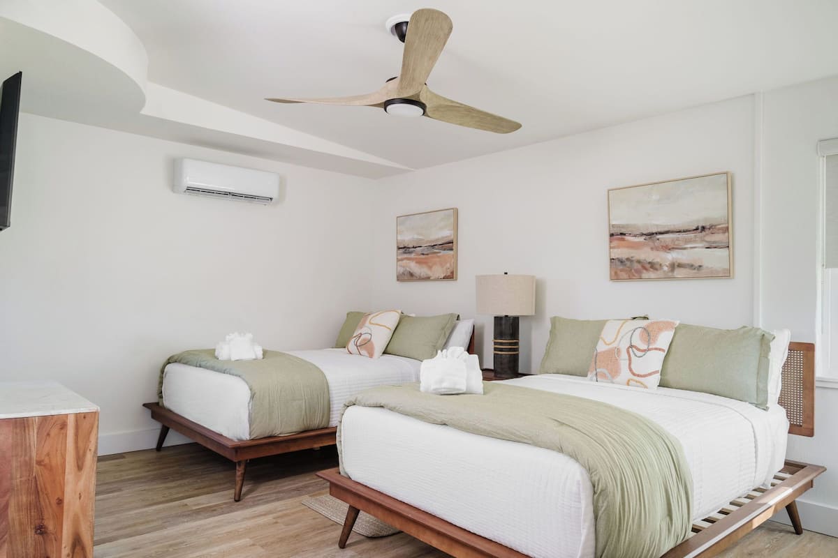 Habitación de hotel con dos camas, ventilador de techo, unidad de aire acondicionado y obras de arte en las paredes.