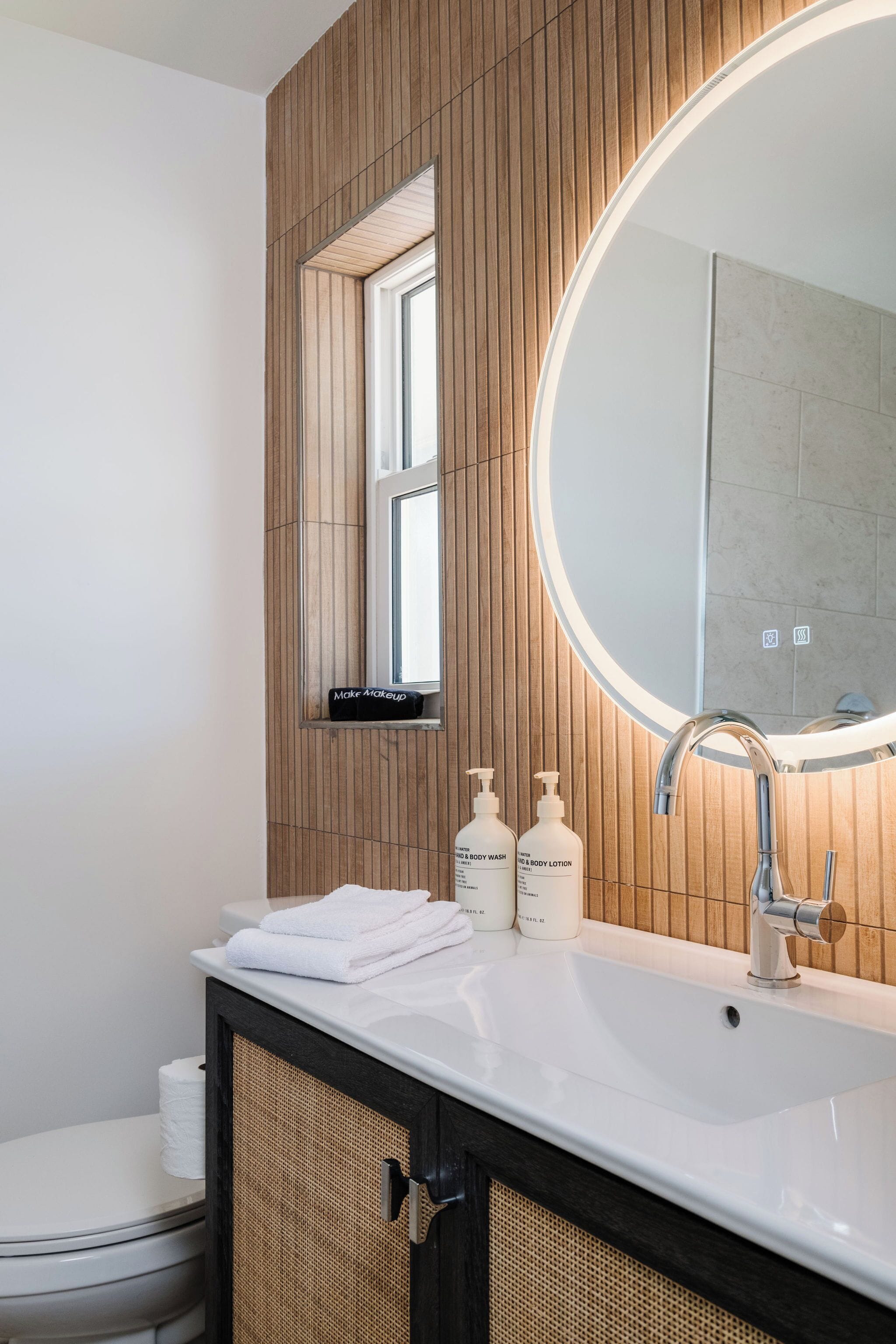 A modern bathroom with a wooden panel wall, a round mirror, a white sink, and two bottles of body lotion.