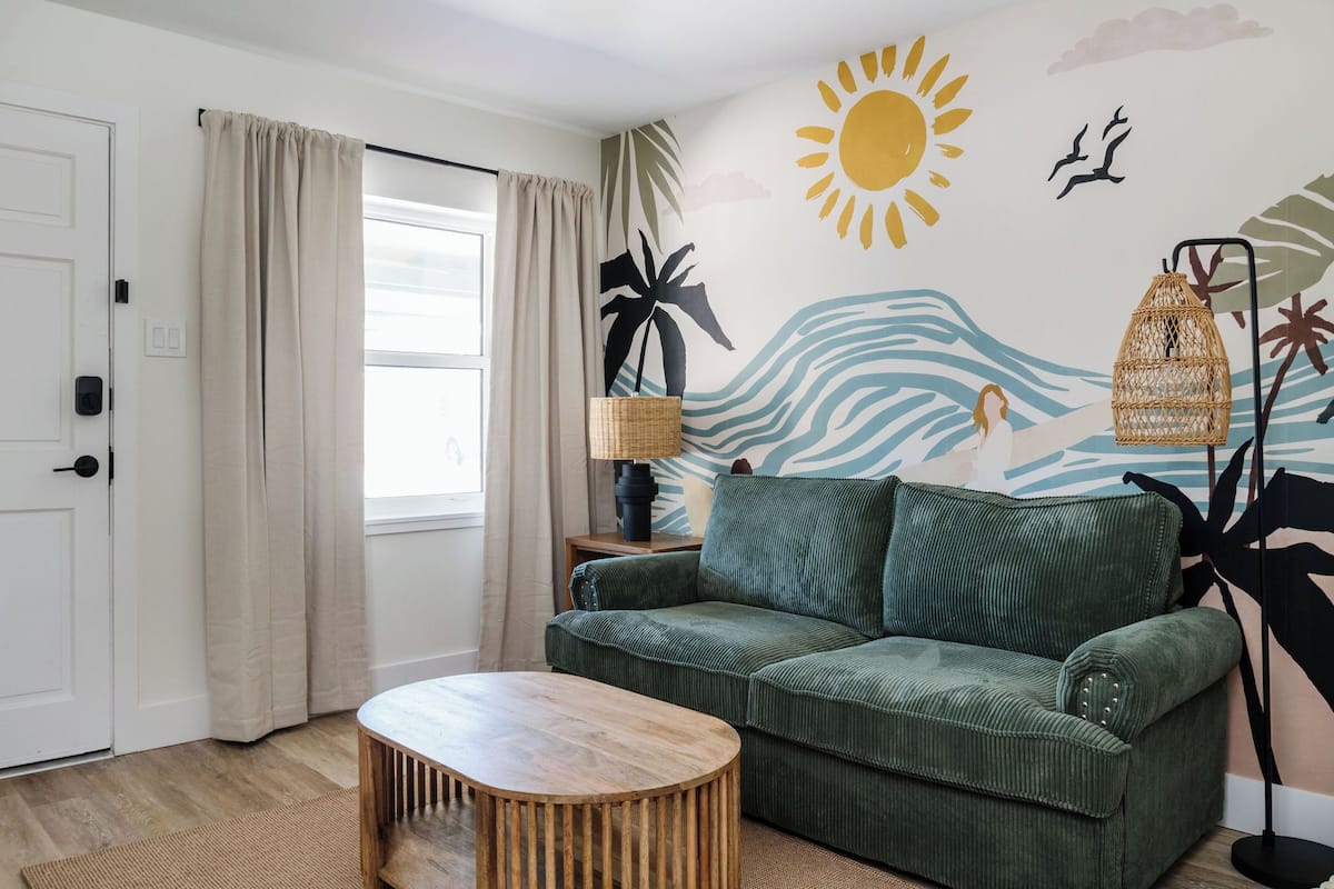 Una habitación con un sofá verde, una mesa de centro de madera, una lámpara y un mural en la pared que representa una escena de playa.