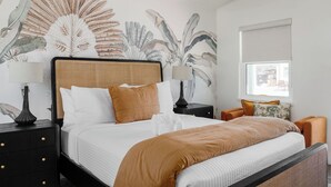 Una cama bien hecha con cabecera, dos mesitas de noche con lámparas, un ventanal con vistas y una pared con un mural de hojas tropicales.