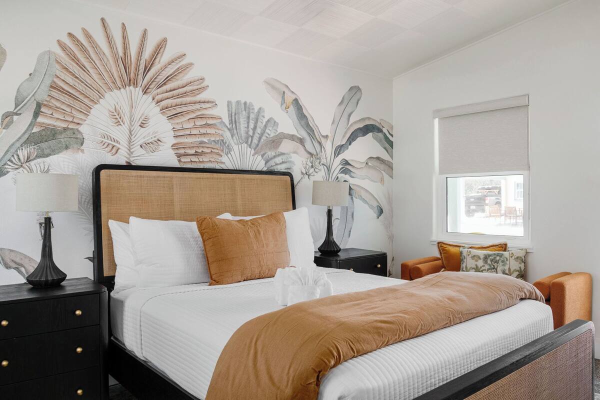 Una cama bien hecha con cabecera, dos mesitas de noche con lámparas, un ventanal con vistas y una pared con un mural de hojas tropicales.