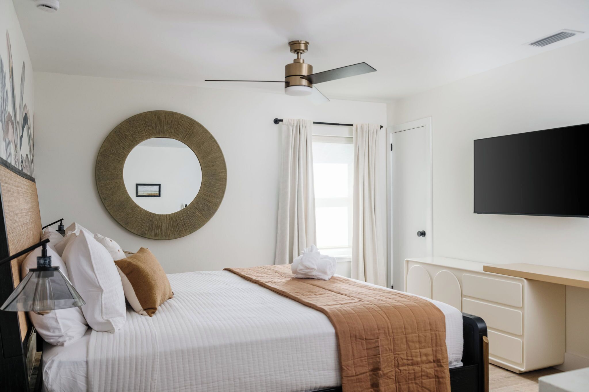 A hotel room with a bed, a round mirror, a ceiling fan, a flat-screen TV, and a desk with drawers.