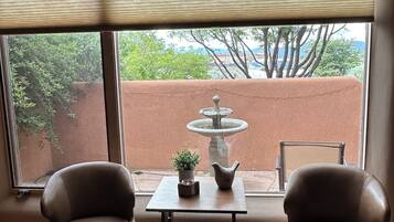 A room with a view of a fountain and trees, two leather chairs, a small table with a plant and a pitcher, and a window with blinds.