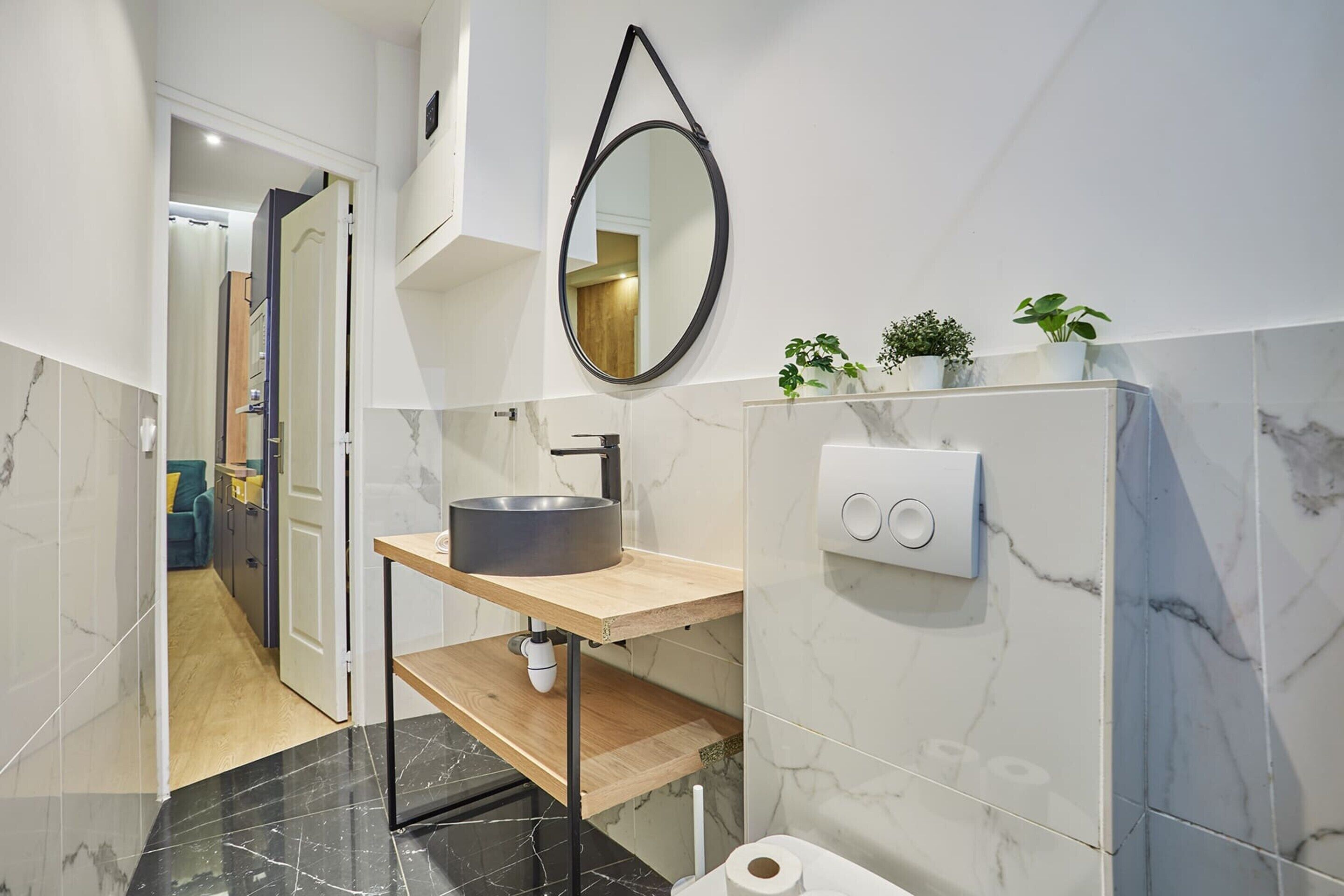 A modern bathroom with a marble floor, a wooden vanity, a round sink, a mirror, and two potted plants.