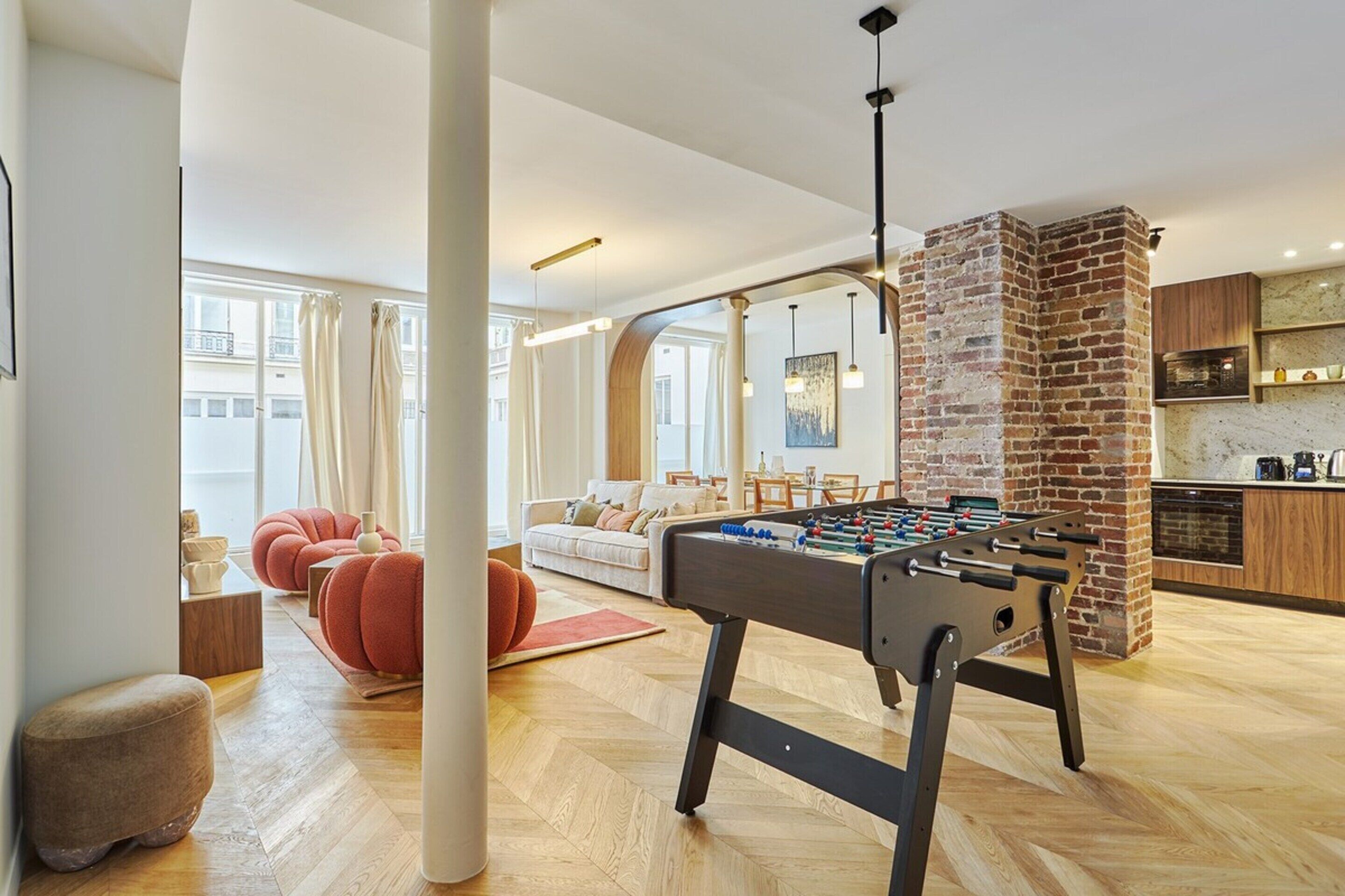 A modern living room with a foosball table, a brick fireplace, and a kitchen area.