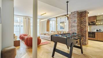 A modern living room with a foosball table, a brick fireplace, and a kitchen area.