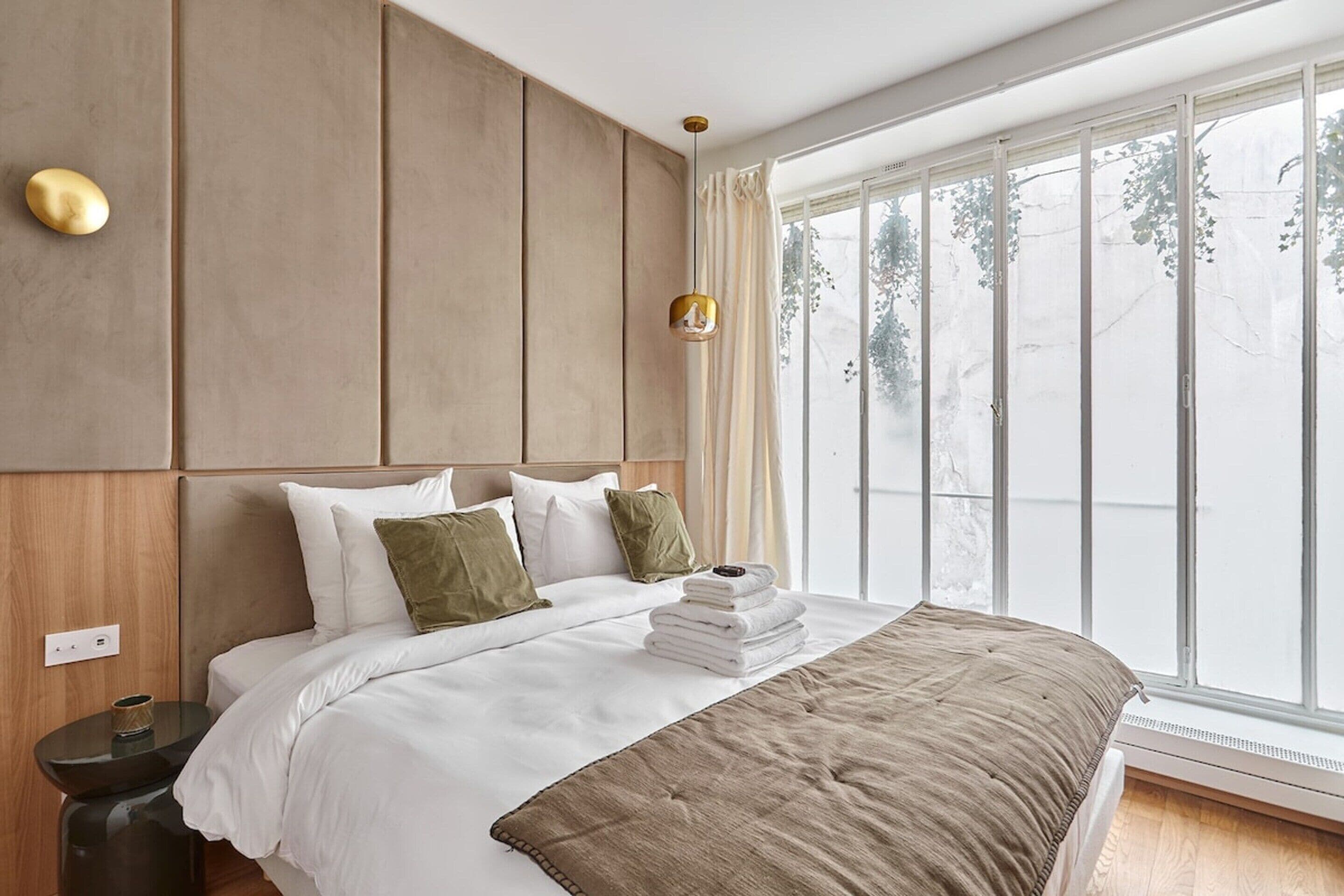A modern bedroom with a large bed, a wooden headboard, a bedside table with a lamp, and a view of trees through a large window.