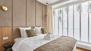 Un dormitorio moderno con una cama grande, cabecera de madera, mesita de noche con lámpara y vista a árboles a través de un ventanal amplio.