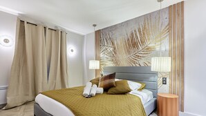 Un letto rifatto con cura, con una testiera grigia, una coperta beige e un cuscino marrone. Accanto al letto, un tavolino rotondo in legno.