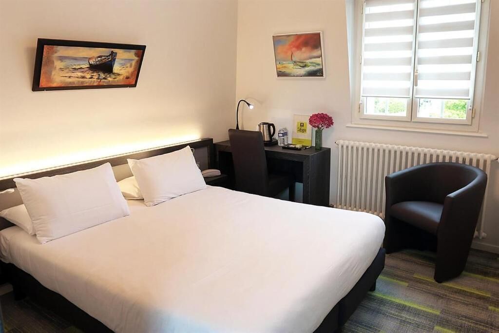A hotel room with a bed, a desk, a chair, and two paintings on the wall.