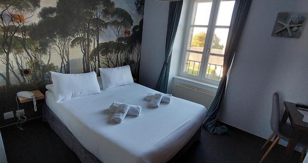A neatly made bed with white linens and towels, a nightstand with a phone, a window with curtains, and a mural of trees on the wall.