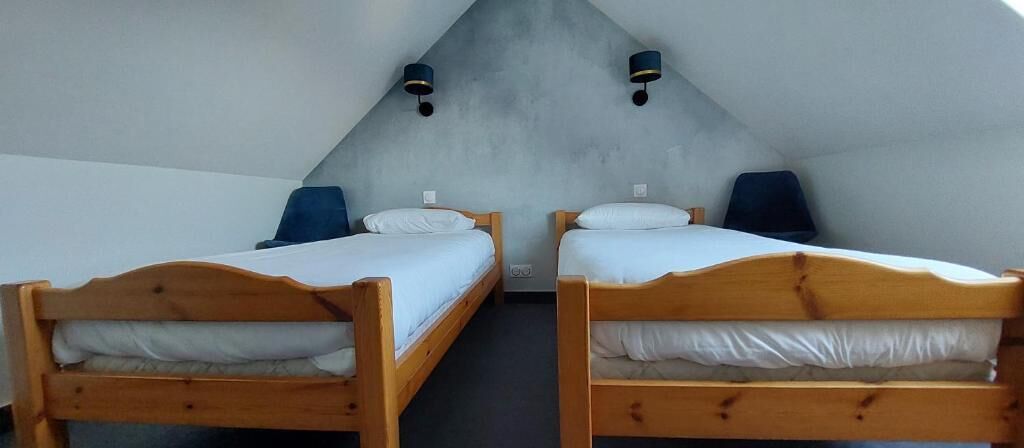 Duas camas de madeira com lençóis brancos em um quarto com teto inclinado e parede em tom claro.