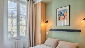 Une chambre d’hôtel avec un lit, une fenêtre avec des rideaux, une lampe fixée au mur et un tableau encadré.