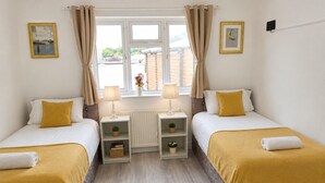 Une chambre double avec deux lits, chacun recouvert d’une couverture jaune, des tables de chevet avec des lampes, un radiateur et une fenêtre avec des rideaux.