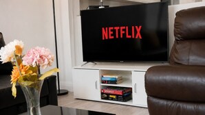 Un salon avec un téléviseur affichant le logo « NETFLIX », un canapé en cuir marron, un meuble blanc contenant des jeux de société et un vase rempli de fleurs posé sur une table.