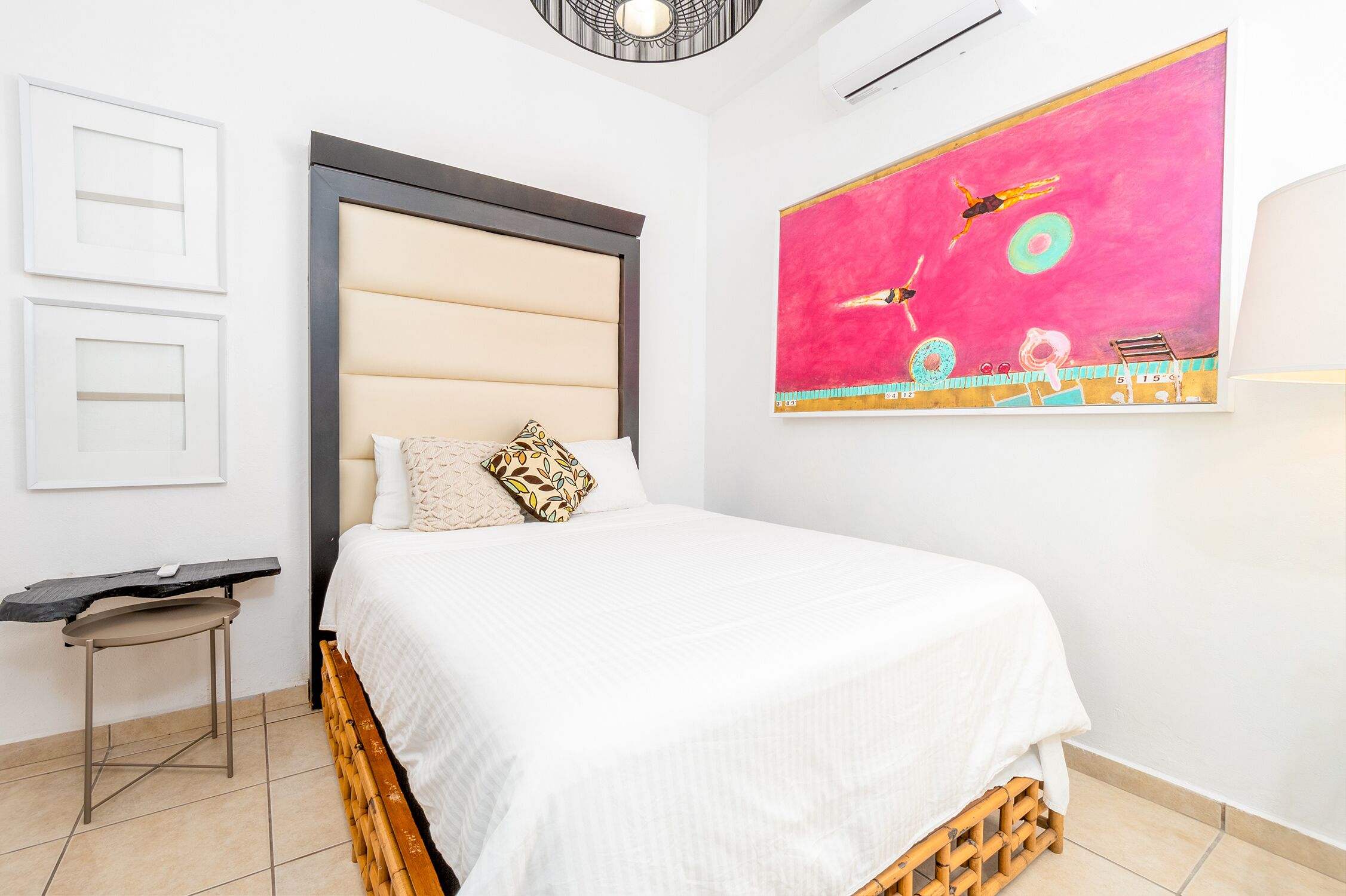 A bedroom with a bed, a small table, a wall-mounted air conditioner, and a large abstract painting.