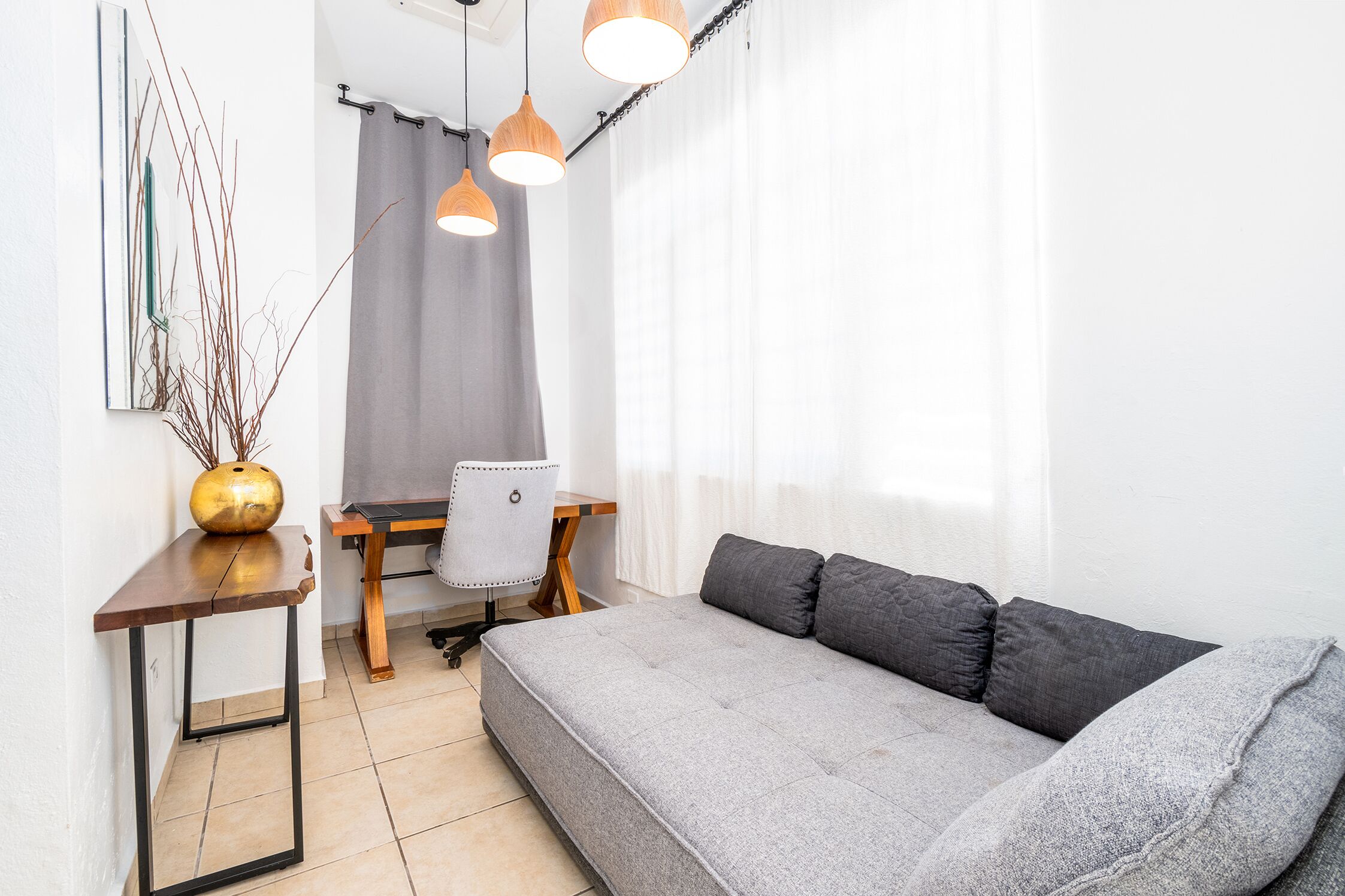 A modern living room with a grey sofa, a small wooden table, and a chair. There are pendant lights above the table and a vase with branches on the table.