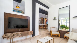 Un soggiorno moderno con una grande TV a schermo piatto, una panca in legno, un tavolino, sedie in vimini e un ampio specchio.