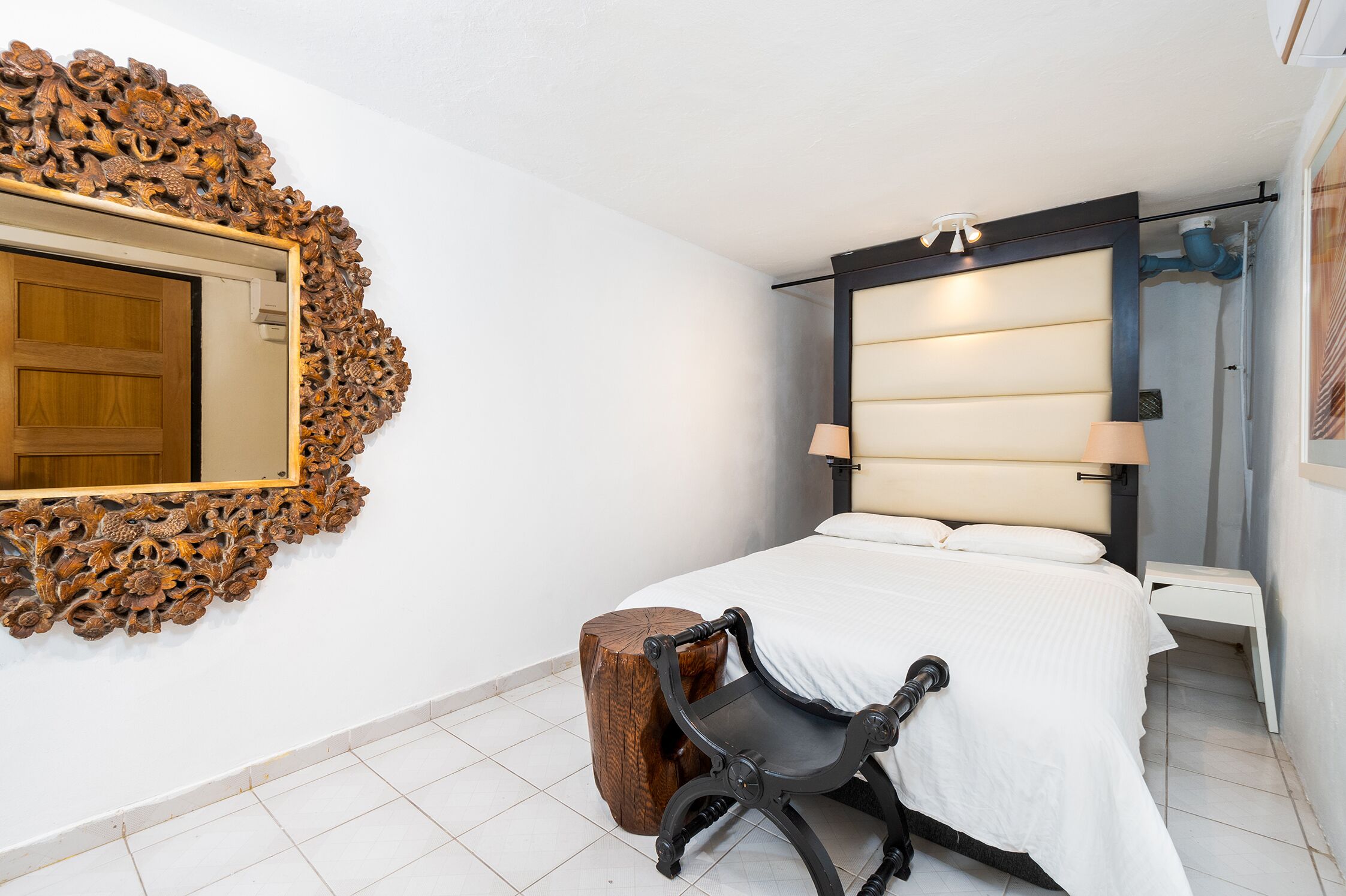 Ein Schlafzimmer mit einem großen Spiegel, einem Bett mit weißer Bettwäsche, einem hölzernen Hocker und einem Nachttisch mit einer Lampe.