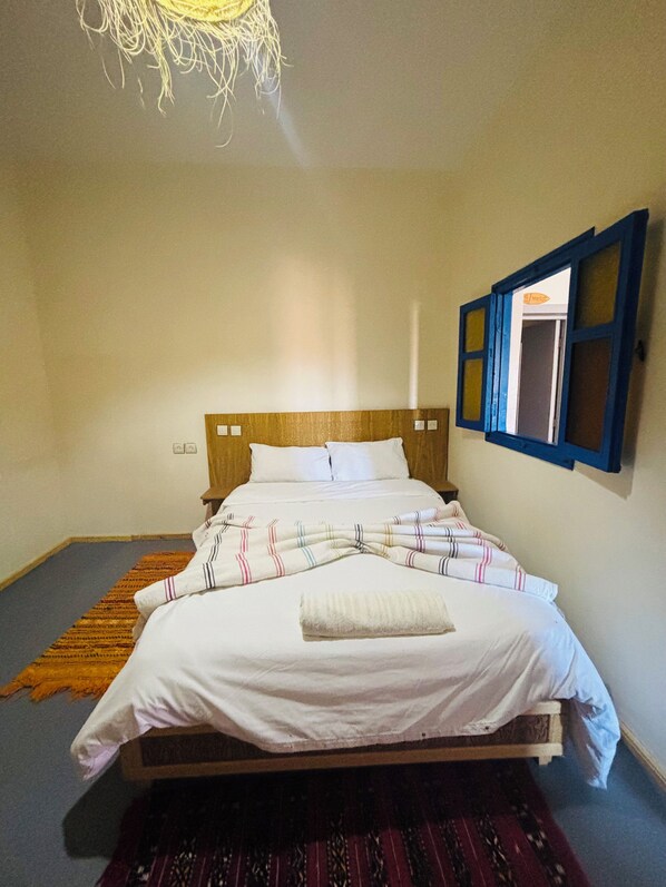 A bed with white linens, a wooden headboard, and a window with blue shutters. - Sunrise Camp Mirleft (Mirleft)