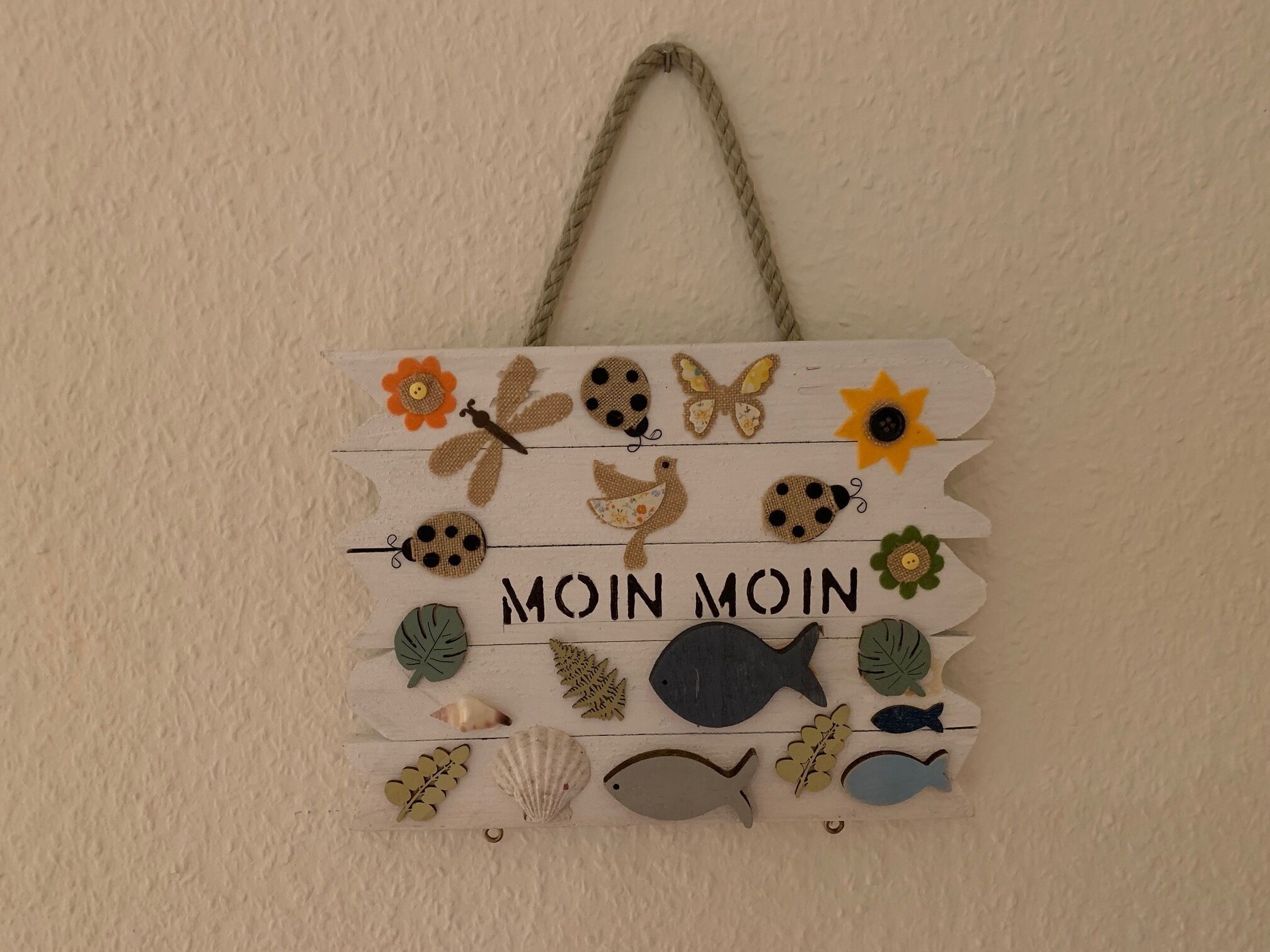 Ein dekoratives Wandbild mit verschiedenen Bastelarbeiten wie Blumen, Schmetterlingen und Fischen, mit dem Text „MOIN MOIN“.