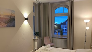 A hotel room with a bed, two bedside tables, a chair, a window with a view of buildings, and wall-mounted lights. - Meeresbrise 1 Top location with garden use. Pet friendly! (Tönning)