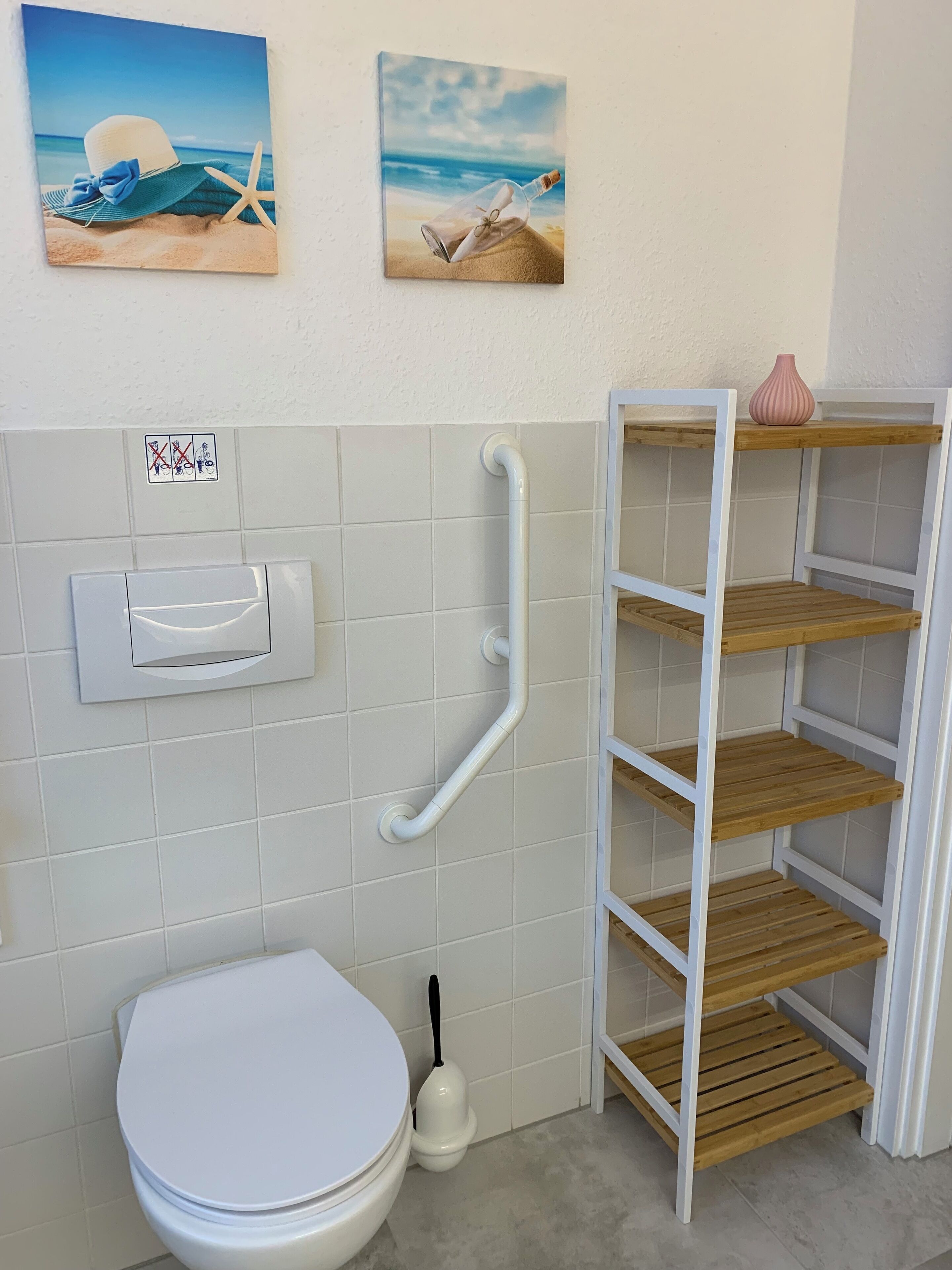 Une salle de bain avec des toilettes blanches, une barre d’appui, une étagère et deux tableaux accrochés au mur.