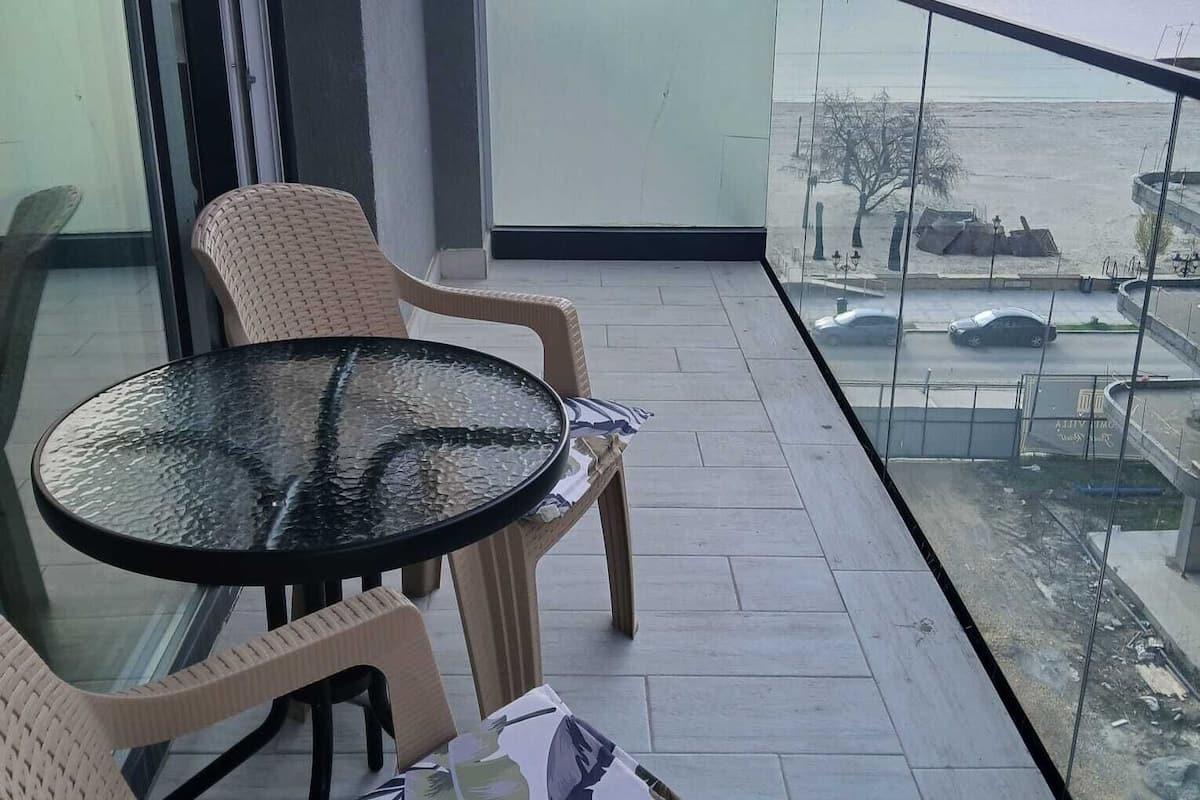 Un balcon avec une table en verre et des chaises en osier, donnant sur une étendue d’eau et un parking.
