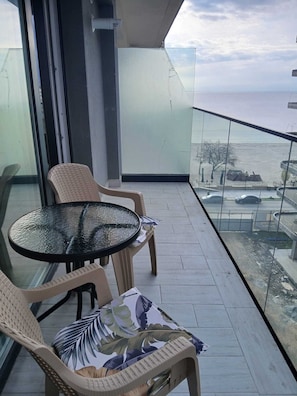 Un balcon avec une table en verre et des chaises en osier, donnant sur une étendue d’eau et un parking.