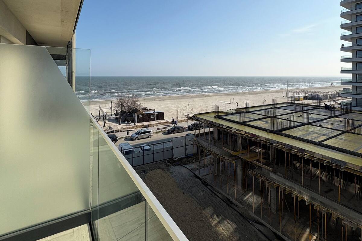 Un bâtiment moderne doté d’un balcon en verre donnant sur une plage et un chantier de construction.
