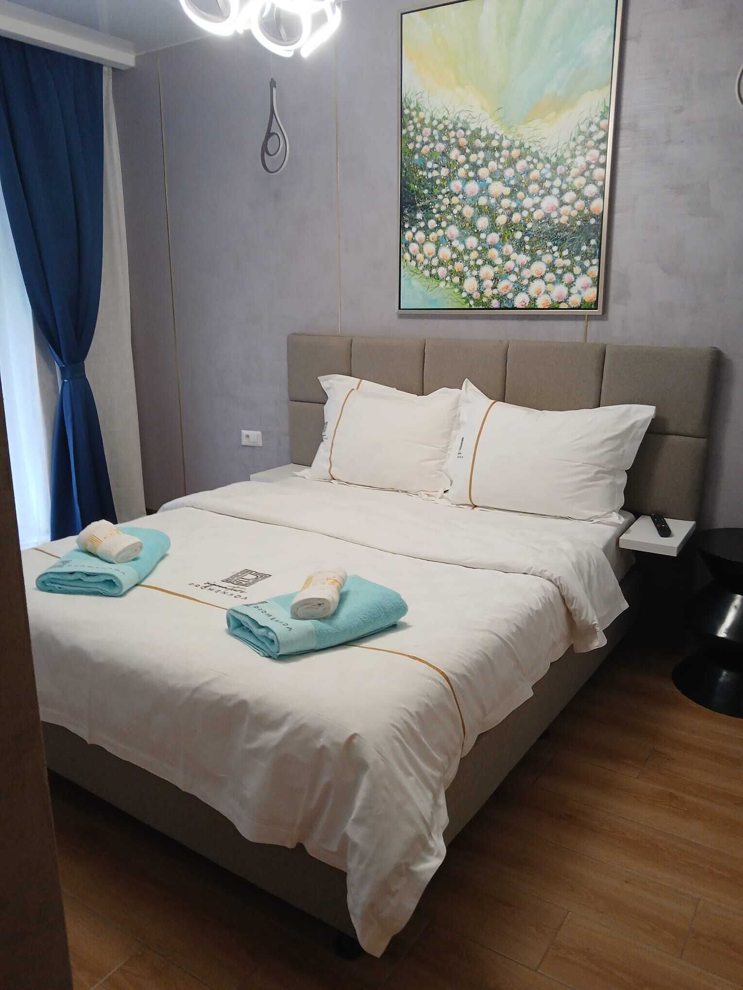Una cama bien hecha con ropa blanca, una toalla azul y una toalla enrollada sobre la cama. Hay un cuadro de flores en la pared encima de la cama.