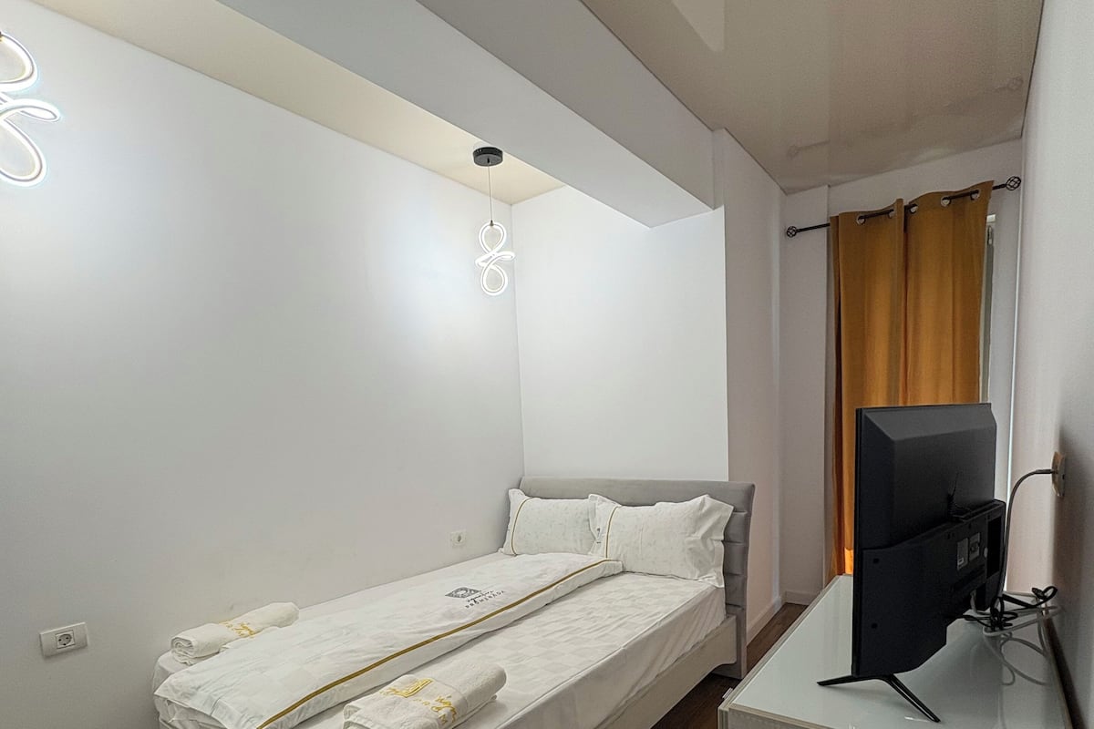 Une chambre de taille modeste, équipée d’un lit simple, d’un téléviseur à écran plat et d’un petit bureau.
