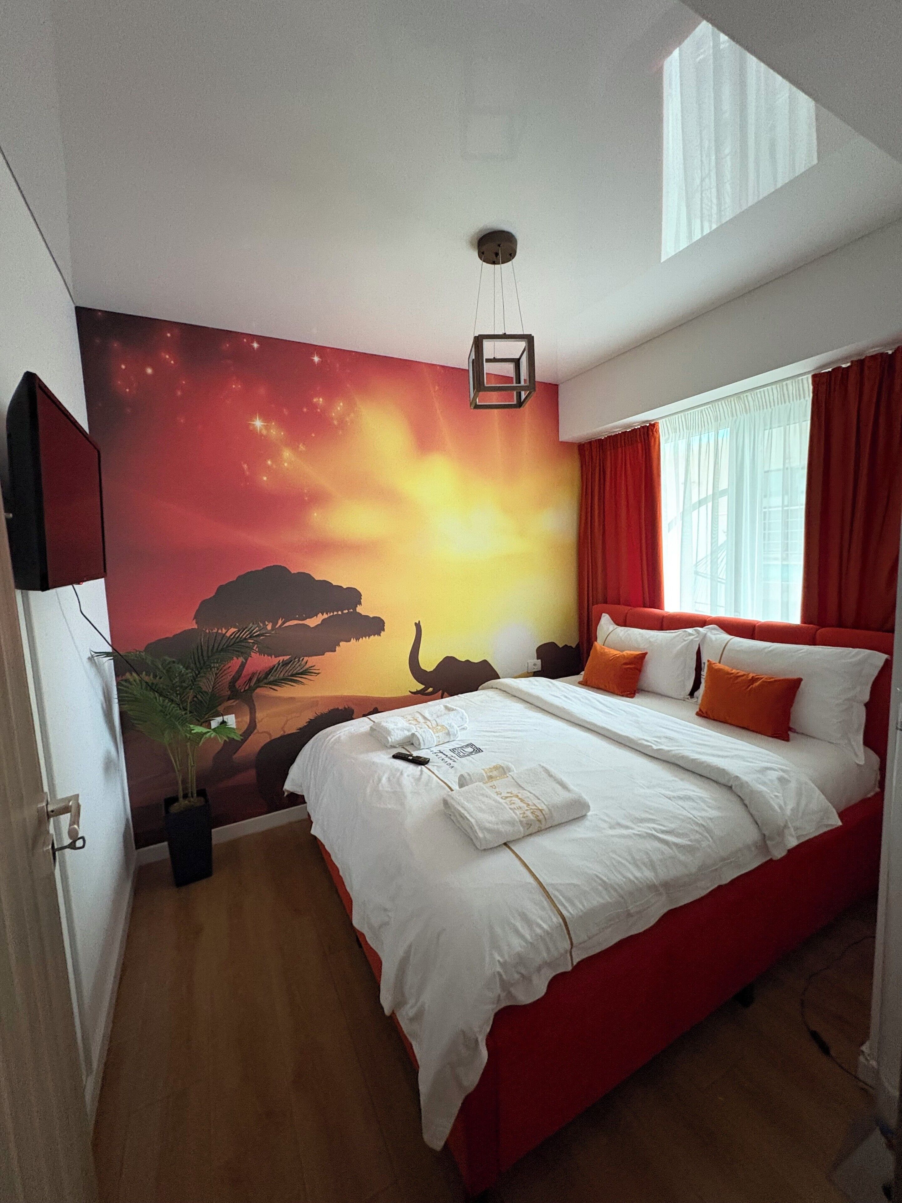 Habitación de hotel con una cama grande, cortinas rojas, una lámpara en el techo, un mural en la pared y una planta en maceta.