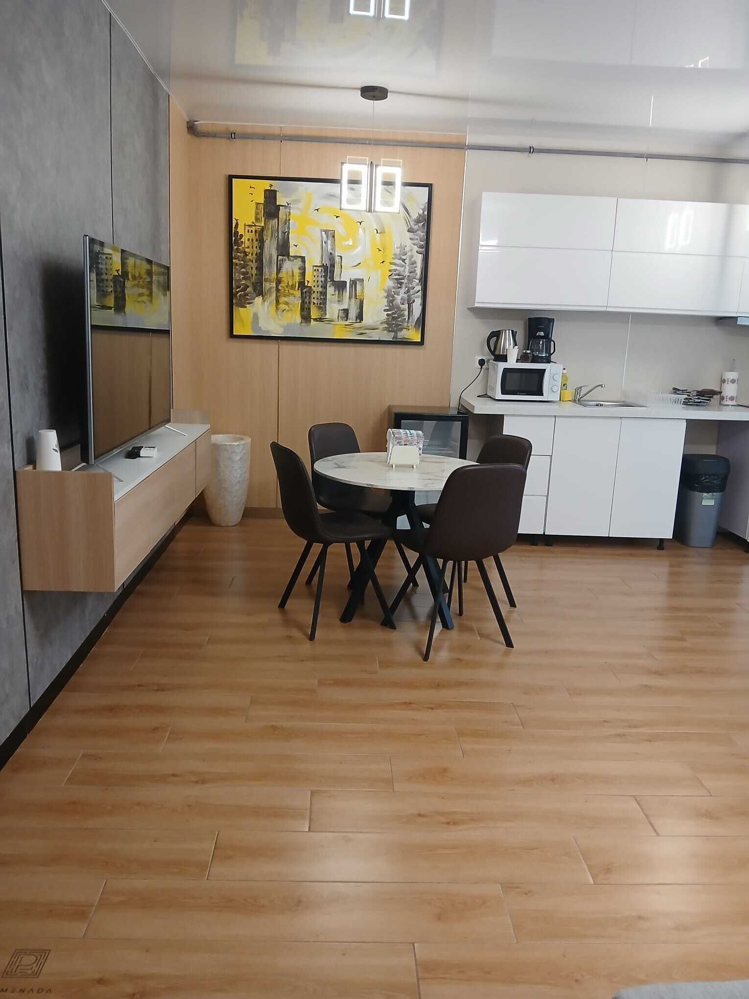 Una cuina moderna amb armaris blancs, una taula de dinar amb caires, un quadre a la paret i un televisor sobre un moble.