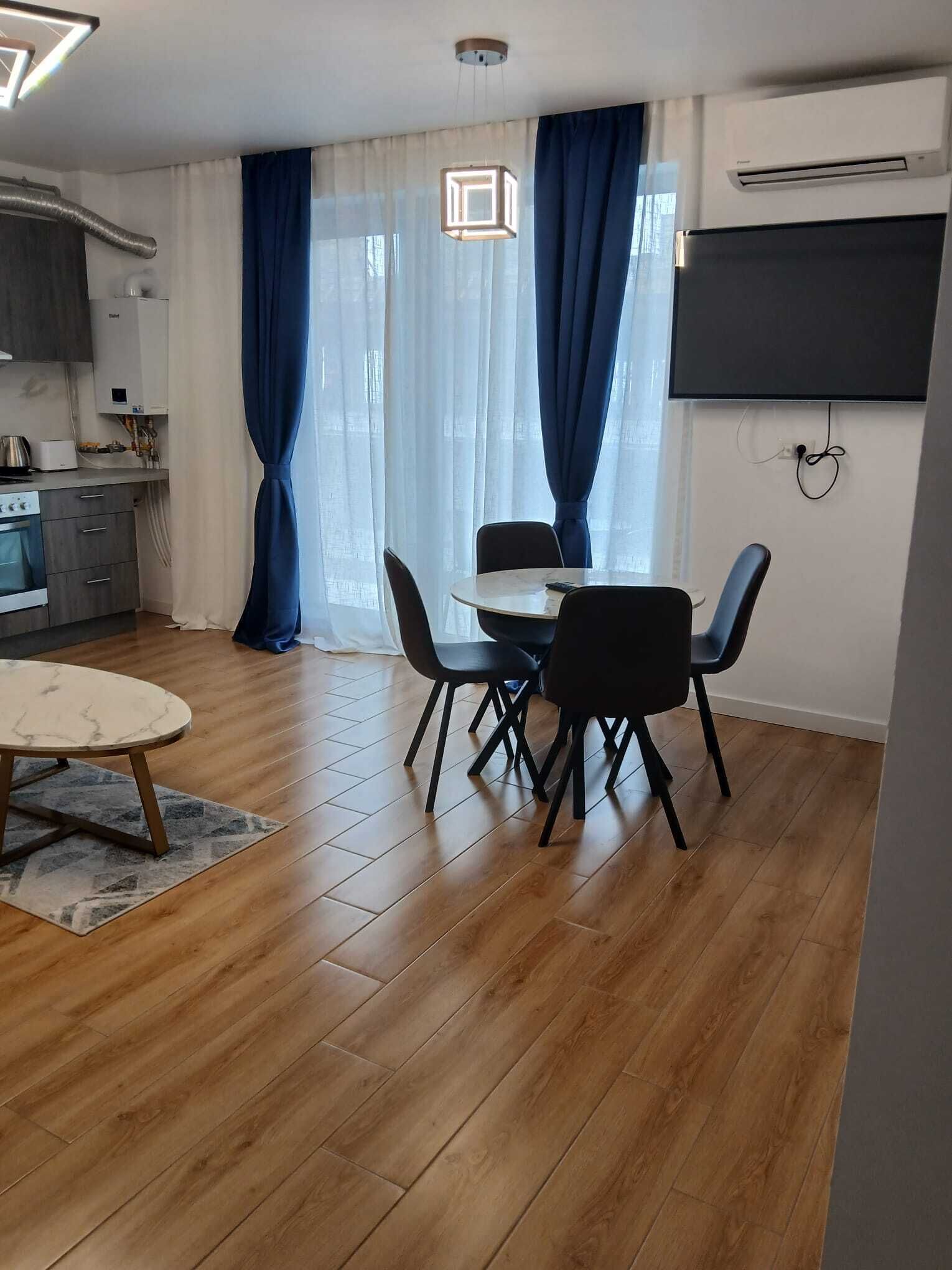 A modern kitchen with a built-in oven, a dining area with a round table and four chairs, and a large window with blue curtains.