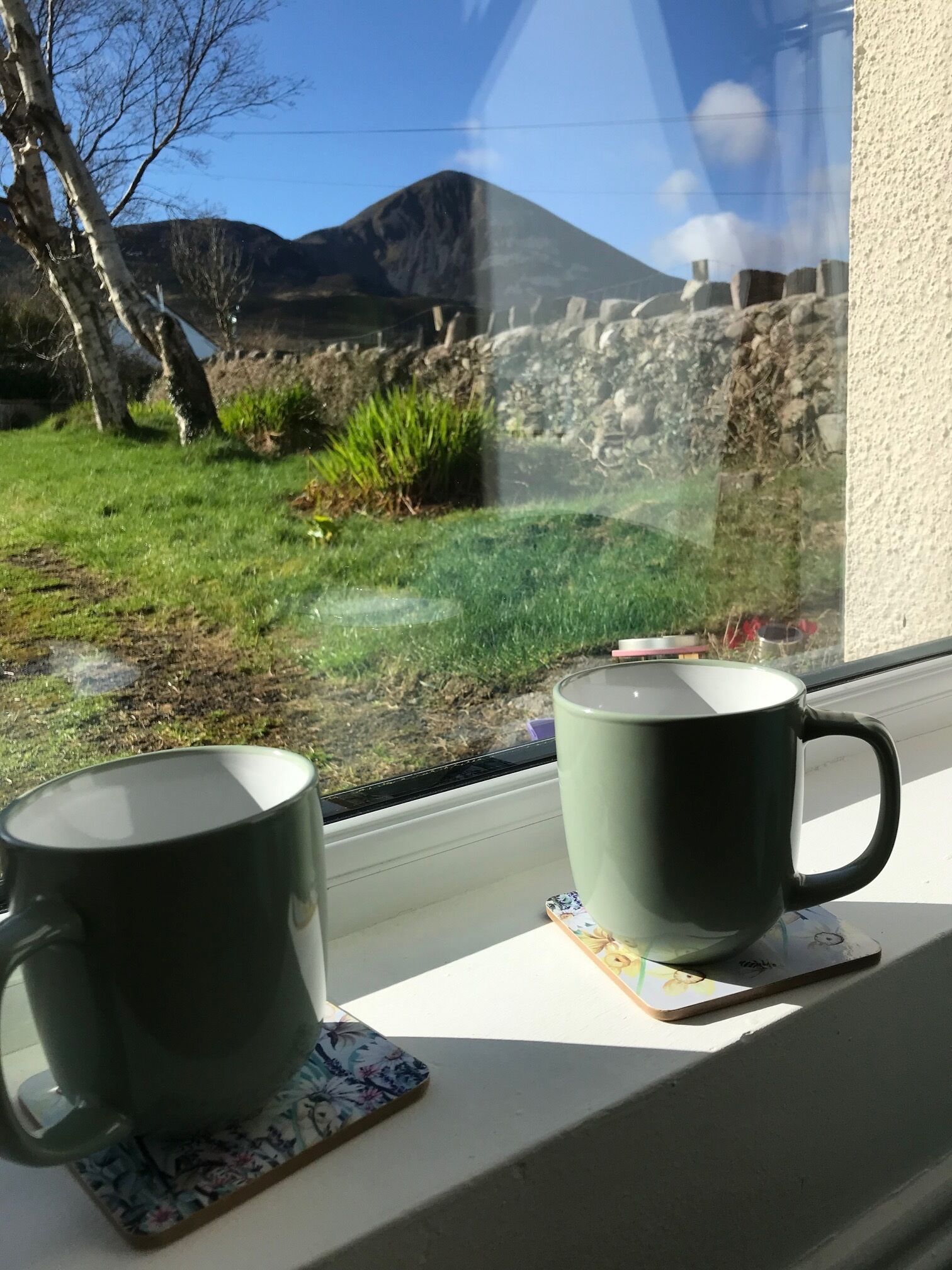 Dos tazas sobre un alféizar con vistas a un campo verde y montañas al fondo.
