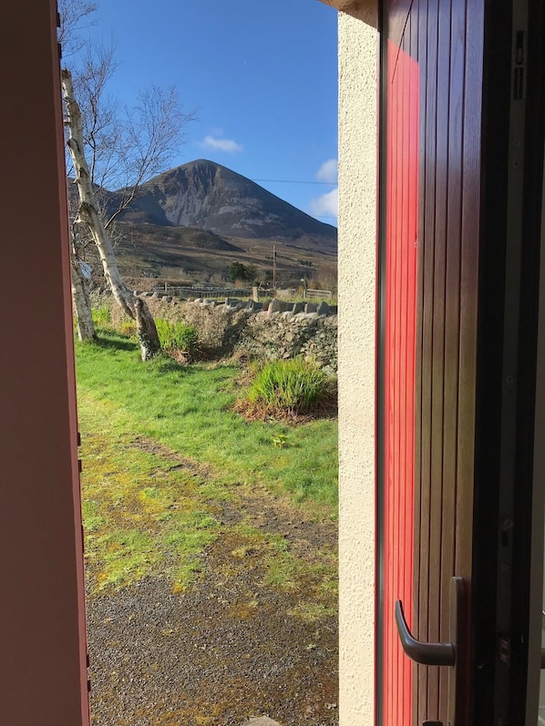 A view from a doorway with a red door, leading to a grassy area with a stone wall and a mountain in the background. - Comfy Annexe near Croagh Patrick with sea views
(Murrisk)