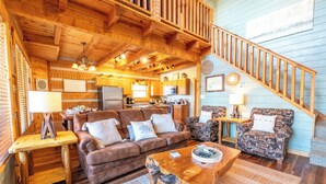 Interior acogedor de una cabaña con techo de madera, una escalera, una sala de estar con sofá marrón y sillones estampados, y un área de cocina.