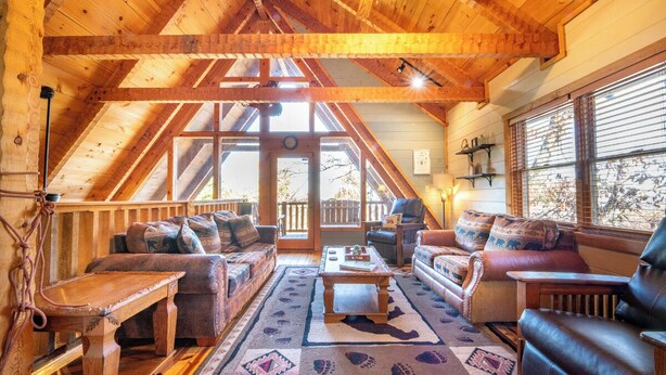 Interior acogedor de una cabaña de troncos con techo alto, vigas de madera, un gran ventanal y una chimenea.