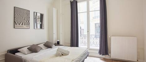 A bedroom with a bed, a radiator, a window with curtains, and two framed pictures on the wall.