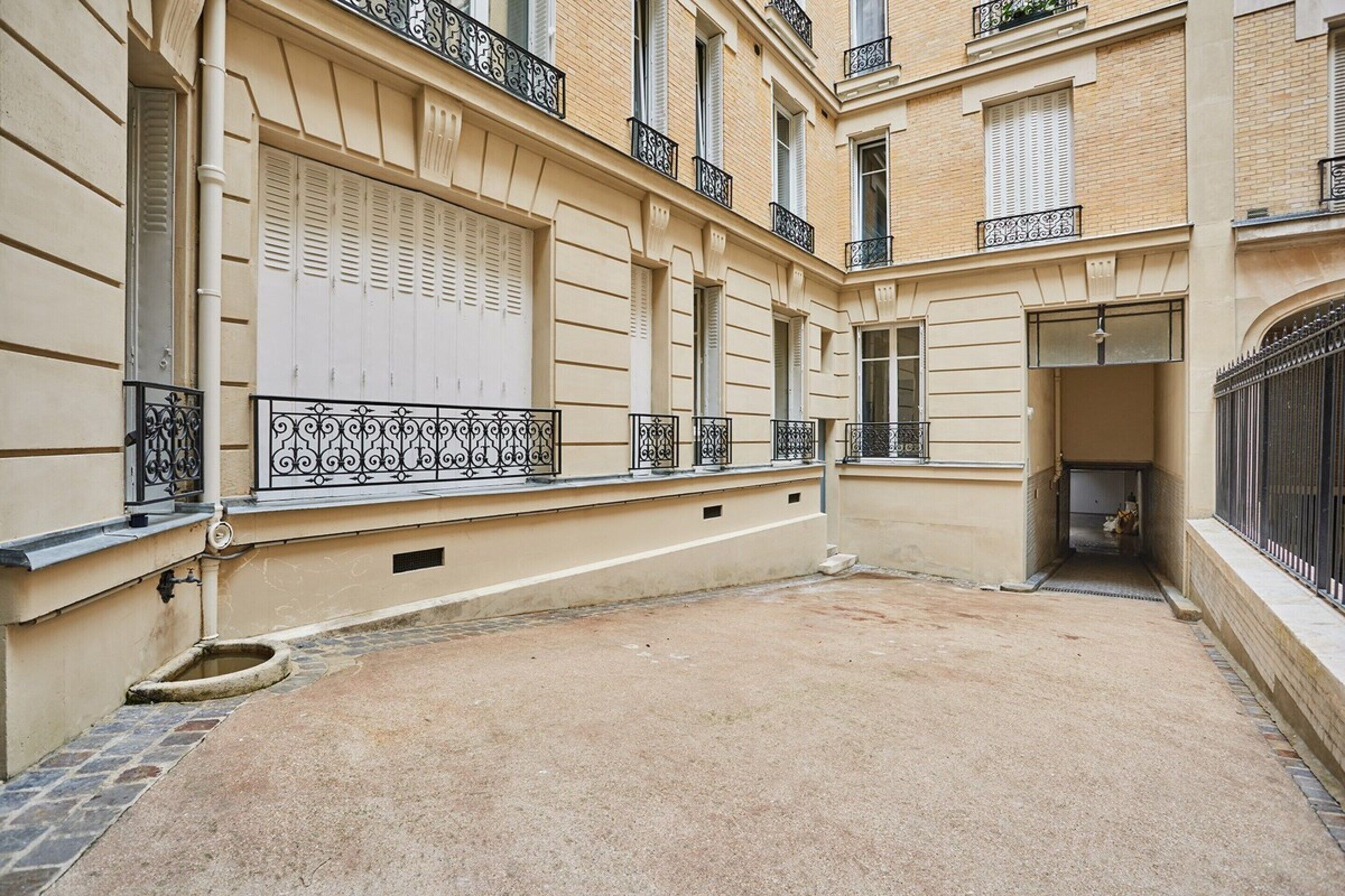 Un patio estrecho con un edificio color beige, contraventanas blancas y barandillas de hierro forjado negro.