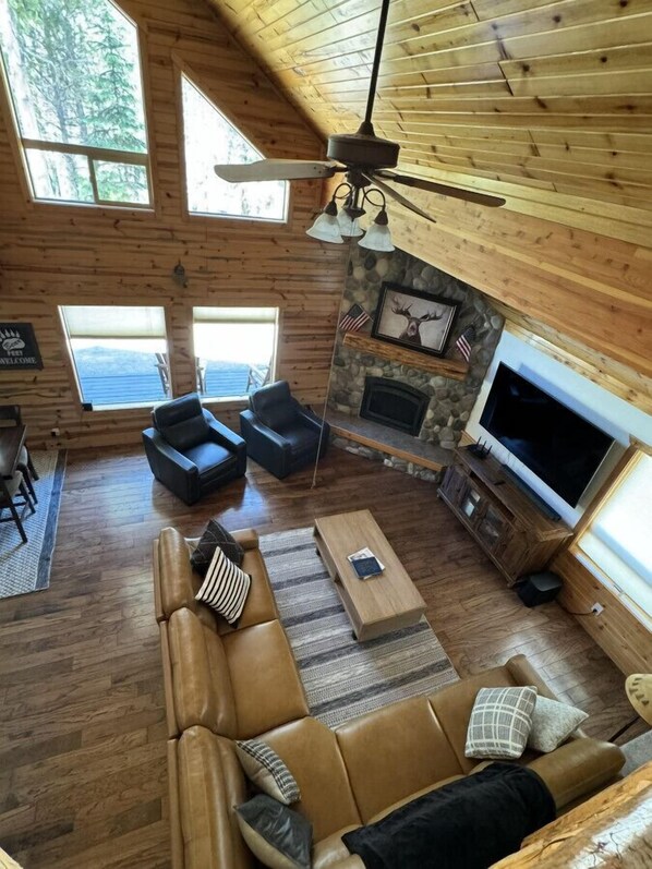 Un salón amplio con chimenea de piedra, sofás de cuero, televisor de pantalla plana y ventanas grandes que ofrecen vistas al exterior.