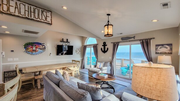 A spacious living room with a view of the ocean, a wooden dining table, and a cozy seating area. - Buccaneers Bounty: Your Oceanfront Treasure Awaits (North Topsail Beach)