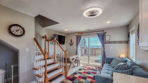 A spacious living room with a wooden staircase, a large window with a view, a blue sofa, and a colorful rug. - Buccaneers Bounty II: Enjoy Stunning Ocean Views (North Topsail Beach)