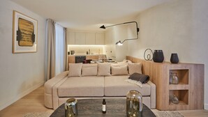 Ein modernes Wohnzimmer mit einem beigen Ecksofa, einer Holzkommode mit Regalen, einem Couchtisch mit Dekoration und einer Küche im Hintergrund.